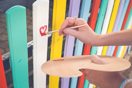 Close up of female hand holding paint brush and drawing red heart shape on colorful wooden fence outdoor. Love and art concepts.