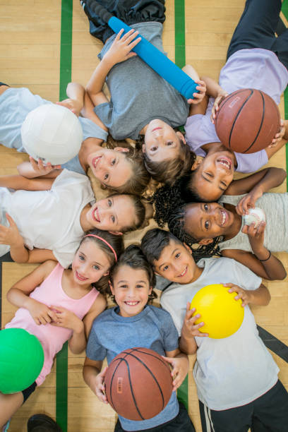 Kids In Gym A multi-ethnic group of children are indoors in a health center. They are wearing casual clothing and running shoes. They are lying on the floor with various sports balls, and smiling up at the camera. community health center stock pictures, royalty-free photos & images