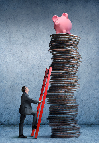 A businessman uses a red ladder as he begins to climb a tall stack of coins that is topped with a pink piggy bank.