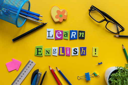 Word LEARN ENGLISH made with carved letters onyellow desk with office or school supplies, stationery. Concept of English language courses.
