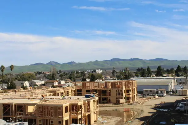Job site in Santa Maria California during framing with green mountains from a fresh rain behind it. Workers pepper the roofs.