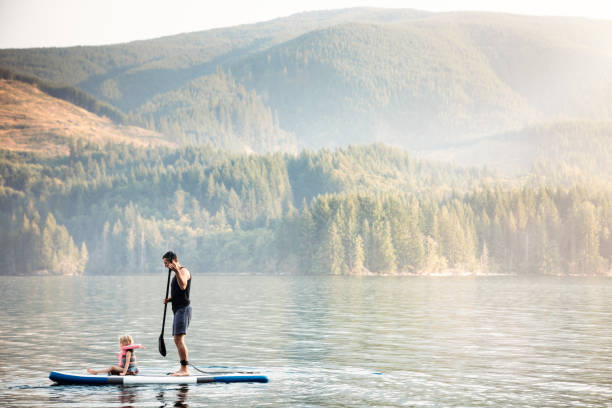 Father and Daughter Standup Paddleboarding A dad paddles on his SUP, his little girl sitting on the front of the board.  A fun time of bonding and togetherness in a beautiful lake setting.  Shot in Cougar, Washington, USA. northwest stock pictures, royalty-free photos & images