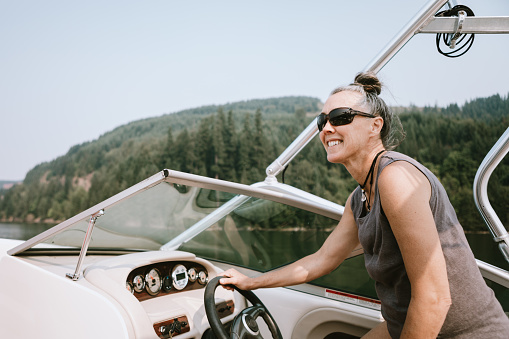 A cheerful woman in her 50's drives a boat on a beautiful mountain lake, a smile of contentment on her face.