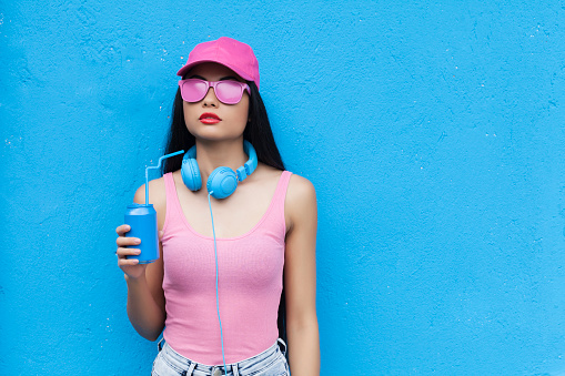 Woman wearing pink vest, blue headphones and holding blue can in front of blue wall background.