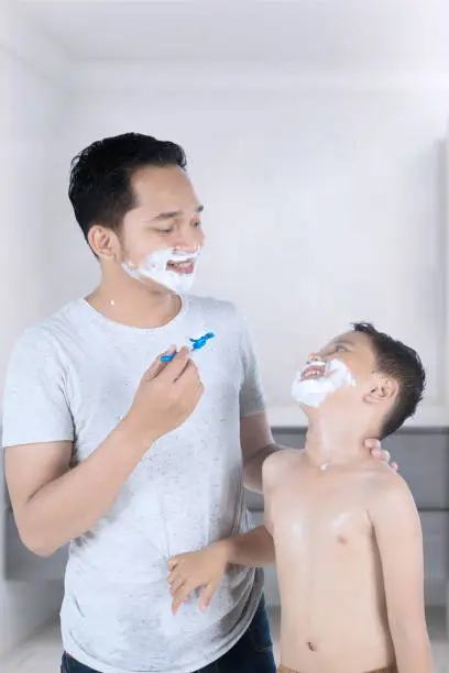 Happy young father and his son using shaving cream to clean and shave their beard in the bathroom