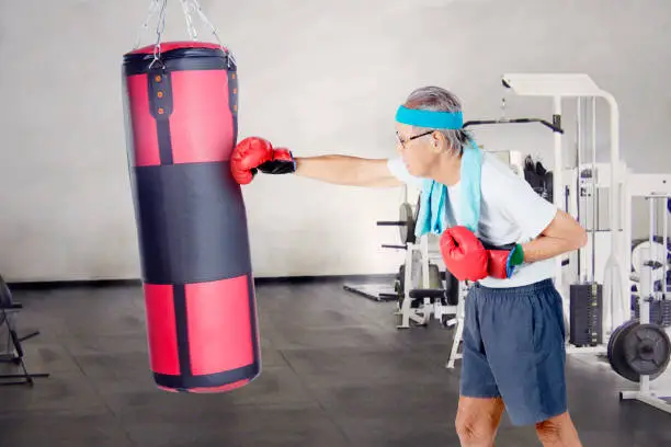 Image of an elderly man practicing boxing by punching a boxing bag in the fitness center