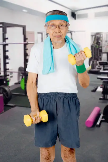 Portrait of an elderly man exercising with two dumbbells and standing in the fitness center