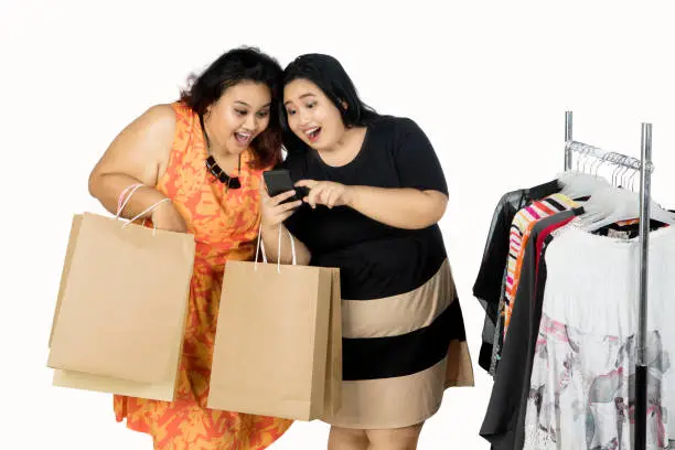 Two young women shopping together while looking at smartphone, isolated on white background