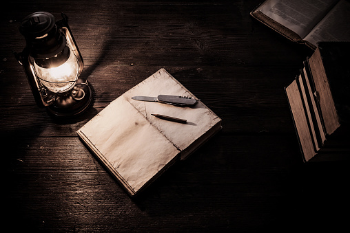 Old-fashioned kerosene lamp and copybook on the dark table in twilight. Soft focus