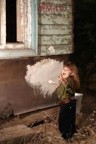 Little caucasian girl dressed as a male colouring a wall of an old destructed house. Laughing and with index finger straight toward camera lens. Construction, repair, education and social concepts.