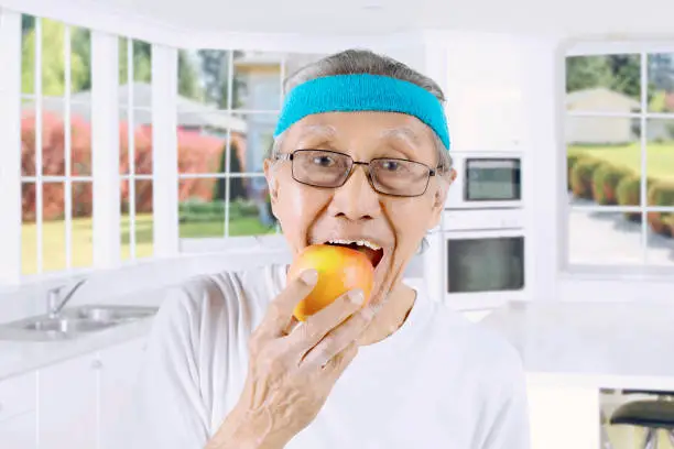 Close up of an elderly man wearing sportswear while eating a fresh apple. Shot in the kitchen