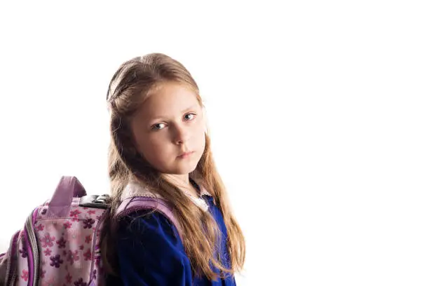 A sad caucasian elementary age schoolgirl posing in uniform and backpack isolated on white background. School and education concept.