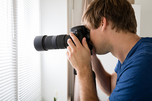 Private Detective Holding Camera Photographing Through Blinds At Home