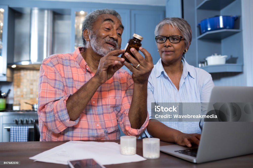 Man looking at medicine while sitting by woman using laptop Man looking at medicine while sitting by woman using laptop in kitchen at home Senior Adult Stock Photo