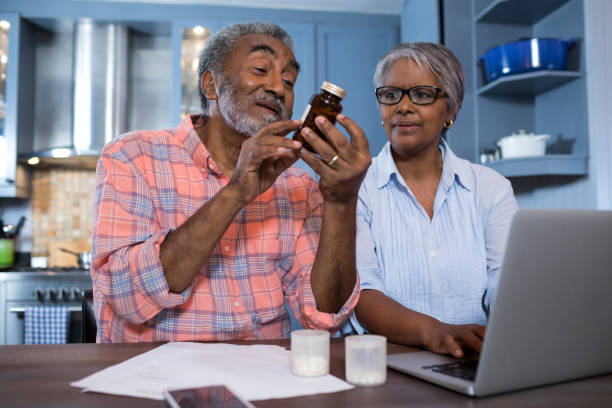 Man looking at medicine while sitting by woman using laptop Man looking at medicine while sitting by woman using laptop in kitchen at home prescription medicine stock pictures, royalty-free photos & images