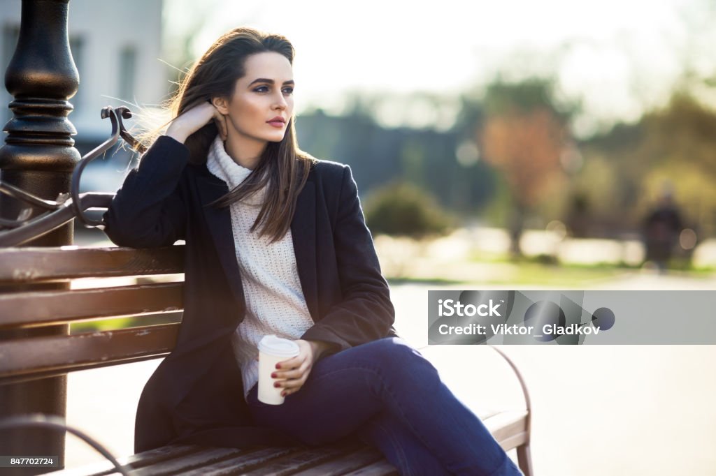 Thoughtful beautiful woman with cup of coffee sitting on a bench in city street Thoughtful beautiful woman with cup of coffee sitting on a bench in city street. Relax concept 20-29 Years Stock Photo