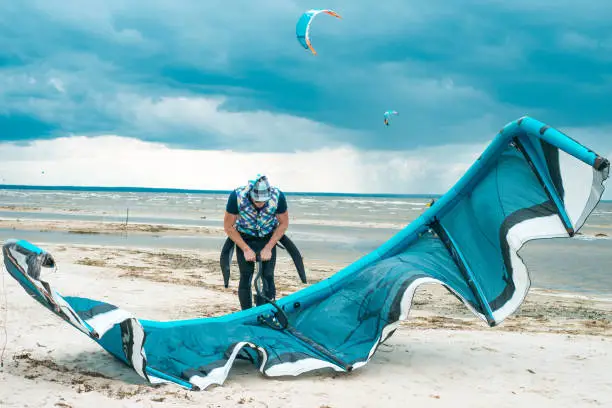 Photo of Kitesurfer pumping fills with air his kite by means of pump with kites in the stormy intense blue sky in the background