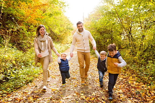 Beautiful young family on a walk in forest. Mother and father with their three sons in warm clothes outside in colorful autumn nature.