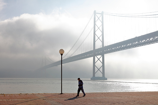 Lisbon, Portugal - January 07, 2015: Lisbon's Ponte 25 de Abril (April 25th bridge) suspension bridge crossing the Tagus river (river Tejo). Man is walking along the promenade close to the river.