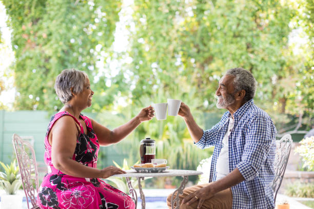 Smiling couple toasting coffee cups while sitting in yard Smiling couple toasting coffee cups while sitting at table in yard older couple drinking coffee stock pictures, royalty-free photos & images