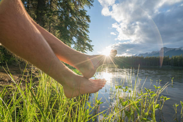 Low angle view of feet dangling from lake pier at sunset, sunbeam and reflection, Canada.