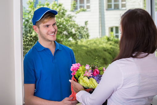Smiling Young Woman Receiving Bouquet Of Flowers From Delivery Man At Home