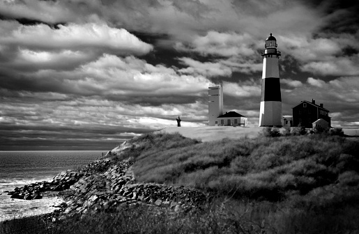 dramatic storm cloud filled sky is backdrop for this picture of the Lighthouse at Montauk Point, Long Island, New York waves crashing against rocks Atlantic Ocean Black and White Image