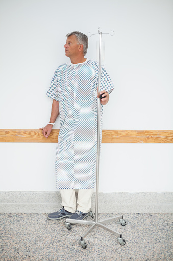 Man is standing against the wall of the corridor in the hospital with his IV drip