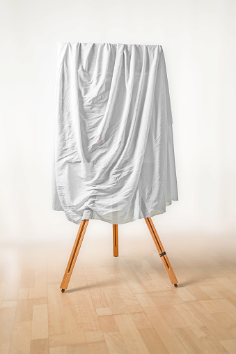 covered painting on an easel, white cloth over the picture, wooden floor and light background, art concept for an exhibition opening day or a presentation ceremony, soft fokus, copy space