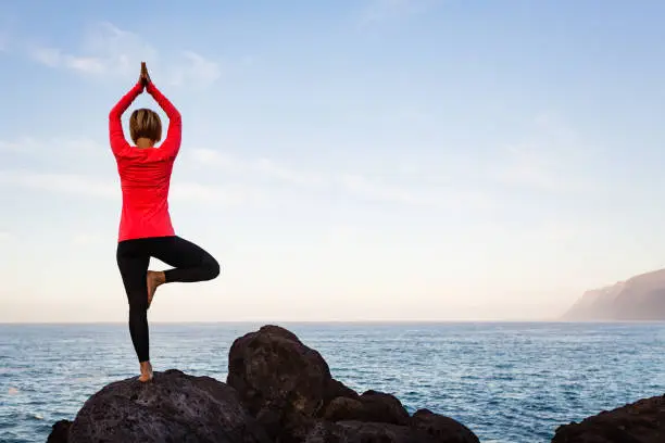 Woman meditating in yoga vrksasana tree pose silhouette at the ocean, beach and rock mountains. Motivation and inspirational fit and exercising. Healthy lifestyle outdoors in nature, fitness concept.