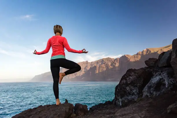 Woman meditating in yoga vrksasana tree pose silhouette at the ocean, beach and rock mountains. Motivation and inspirational fit and exercising. Healthy lifestyle outdoors in nature, fitness concept.