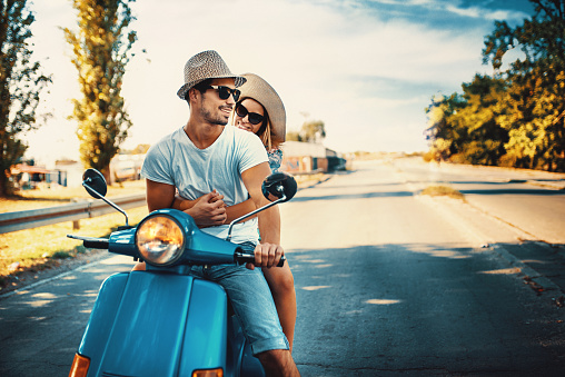 Closeup front view of mid 20's couple cruising a scooter bike on a summer afternoon. They have stopped at red traffic light and posing for the camera.