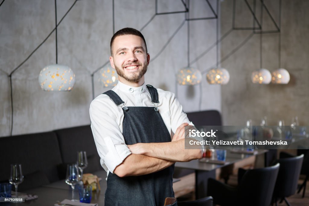 Um garçom em um restaurante, ao lado de mesas com um copo de vinho jovem, sorridente. Ele veste um avental, parece com confiança, braços cruzados sobre o peito - Foto de stock de Restaurante royalty-free