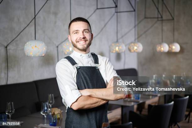 Foto de Um Garçom Em Um Restaurante Ao Lado De Mesas Com Um Copo De Vinho Jovem Sorridente Ele Veste Um Avental Parece Com Confiança Braços Cruzados Sobre O Peito e mais fotos de stock de Restaurante