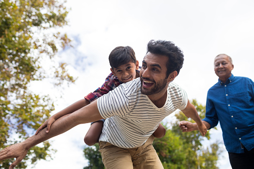 Happy grandfather looking at man giving piggy backing to son with arms oustretched in yard
