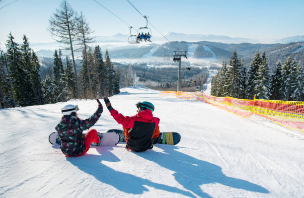 Photo of Snowboarders sit on the top of the ski slope under the ski lift let's high five to each other with a beautiful scenery of mountains and forests on a sunny morning