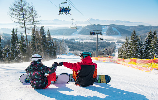 Snowboarders resting on the top of the ski slope under the ski lift at winter resort with a beautiful scenery of the Carpathian mountains and forests on a sunny day