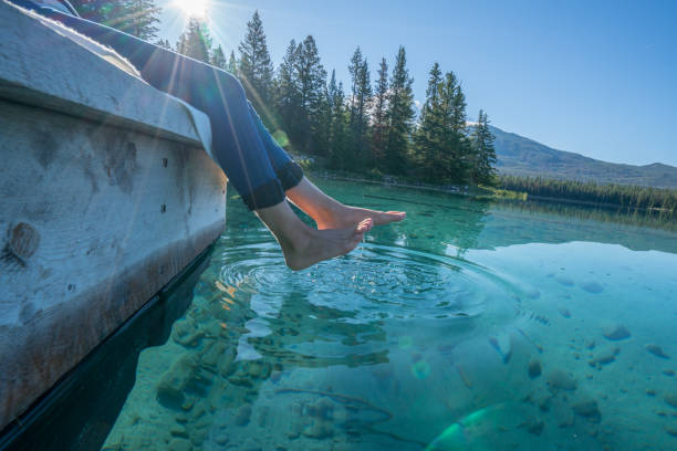 Woman's feet dangle from wooden wharf, above turquoise water lake