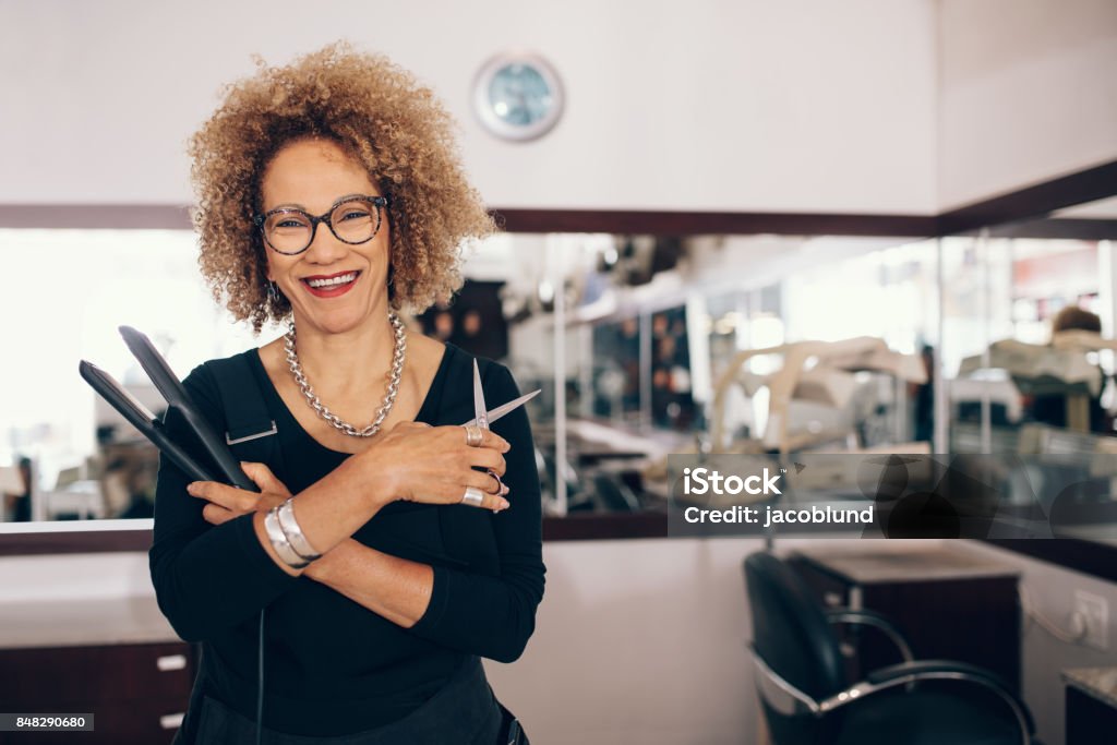 Female hairdresser at the salon holding hairdressing tools Professional hair stylist holding a hair straightener and scissors. Woman hairdresser in happy mood at the salon. Hairdresser Stock Photo