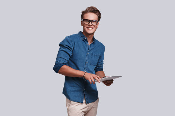 May I help you? Handsome young man looking at camera and holding digital tablet while standing against grey background only young men stock pictures, royalty-free photos & images