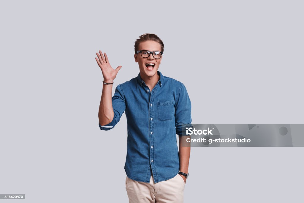 Hey! Good looking young man waving and smiling while standing against grey background Men Stock Photo