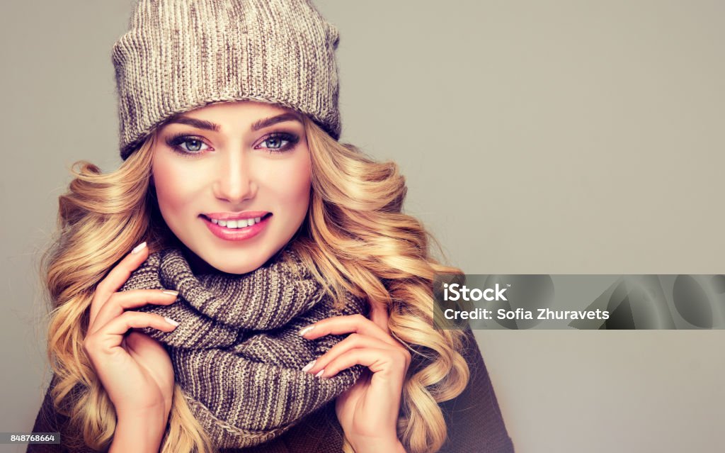 Attractive blonde haired model dressed in stylish knitted winter clothes. Nice, young,smiling blonde woman dressed in gray wool winter knitted hat and scarf. Attractive blonde haired model dressed in stylish winter clothes, hat and snood. Winter Stock Photo