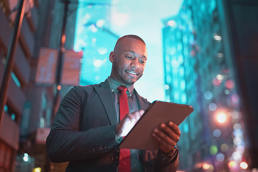 African american businessman looking into his digital tablet reading messages standing in the streets of downtown chicago at night, smiling happy. Edited Colors, Downtown Chicago, Illinois, USA