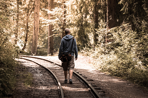 Close up rear view color image depicting a young adult man walking on a deserted railway track in the middle of the forest. He is heading further into the forest, on some kind of journey or adventure. He is wearing a blue hooded top and beige cargo shorts. A black messenger bag or shoulder bag is slung across his back. The railroad track recedes into the distance and curves round a bend. Lots of room for copy space.