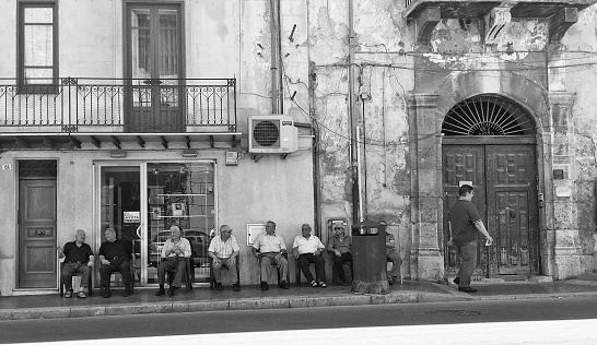 Alcamo, Italy - July 13, 2017: several senior men are seated in a group on the sidewalkof a small Sicilian town's high street, \