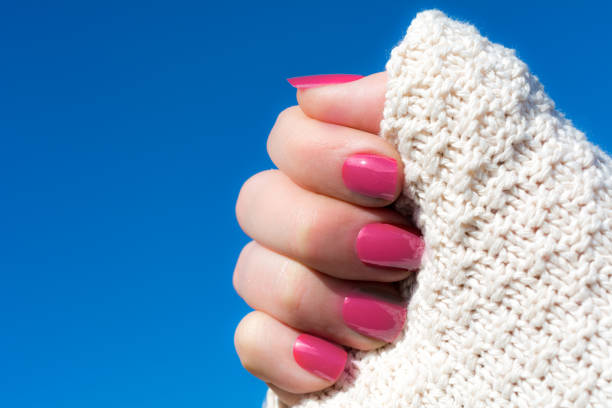 Woman in a white knitted cardigan  showing her short pink manicure against blue sky