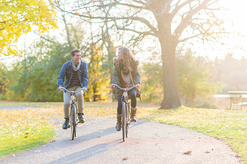 A Caucasian couple are outdoors on a sunny autumn day. They are wearing warm clothes. They are riding bikes on a trail together.