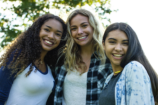 A multi-ethnic group of teenage girls are spending time together in a park at sunset during the summer. They smile and laugh together.