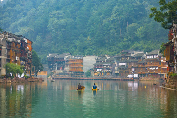 Fenghuang Village Riverside Traditional Houses Personal boats paddling down the center of Tuojiang River lined by traditional wooden houses and forest in Fenghuang, China fenghuang county photos stock pictures, royalty-free photos & images