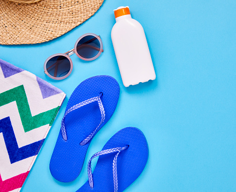 Overhead shot of flip-flop sunglasses suntan lotion sun hat and beach towel. Flat lay shot of summer vacation accessories with copy space. They are color blocked and arranged on blue background.